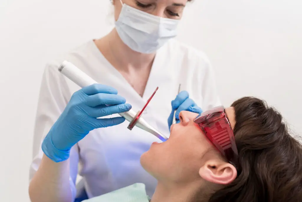 Dentist performing professional teeth whitening on a patient wearing protective red glasses in a dental office.