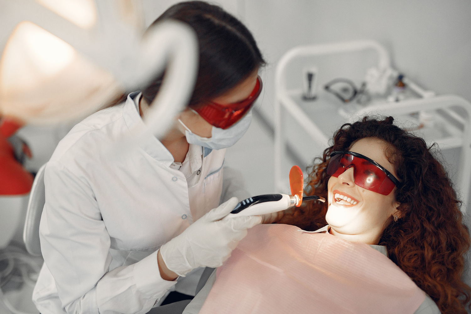 Dentist performing professional teeth whitening on a smiling patient wearing protective red glasses in a dental office.